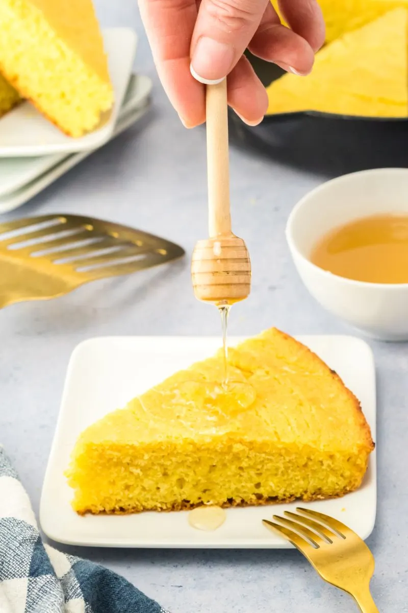 A honey spoon drizzling honey over a wedge of cornbread - Hostess At Heart