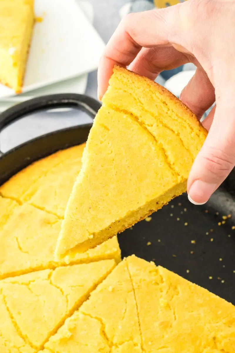 A hand holding a slice of cornbread over a cast iron skillet filled with baked cornbread. Hostess At Heart