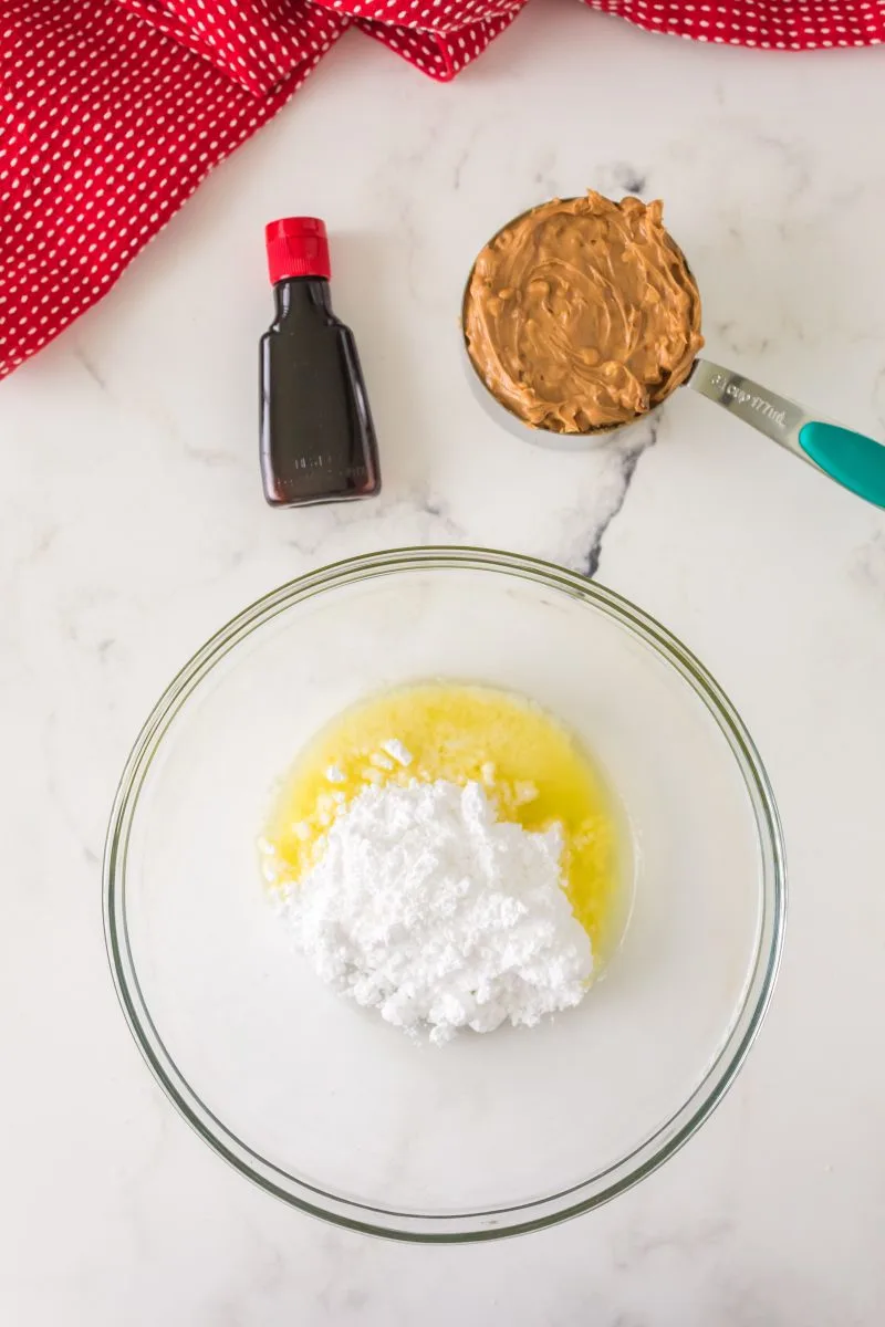 A bowl filled with melted butter and powdered sugar next to a bottle of vanilla extract and a measuring cup filled with chunky peanut butter - Hostess At Heart