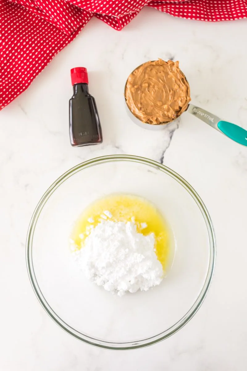 A bowl filled with melted butter and powdered sugar next to a bottle of vanilla extract and a measuring cup filled with chunky peanut butter - Hostess At Heart