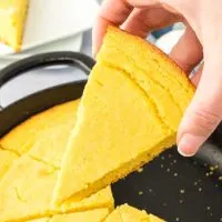 A hand holding a slice of cornbread over a cast iron skillet filled with baked cornbread. Hostess At Heart