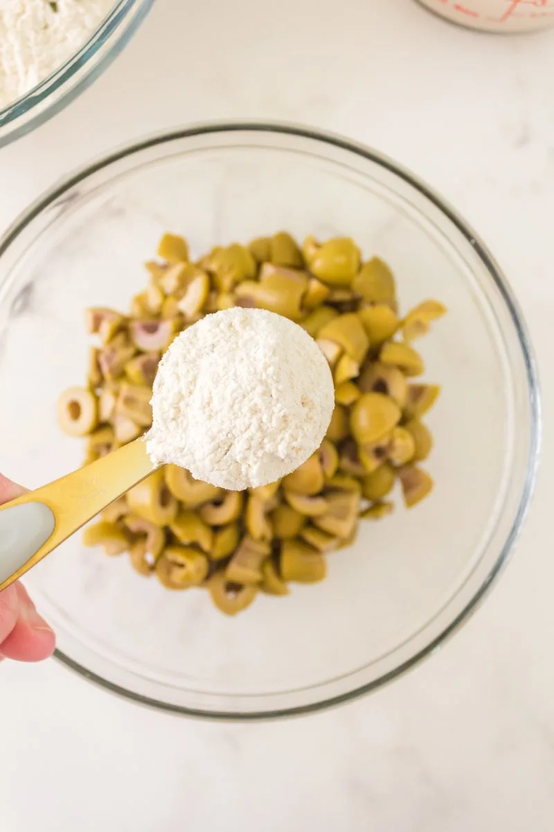A measuring cup of flour hovering over a bowl of chopped green olives - Hostess At Heart