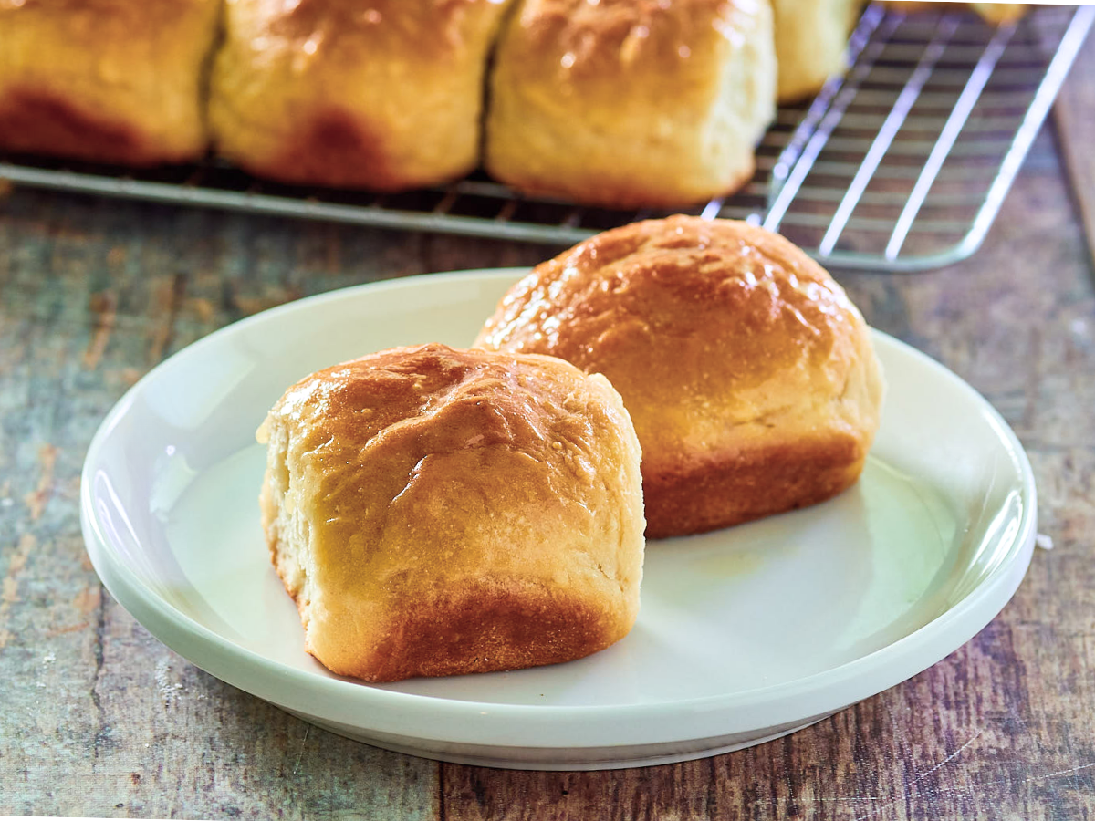 Sideview of two Hawaiian Bread Rolls on a plate. More sweet bread rolls are in the back on a cooling rack. Hostess At Heart