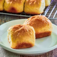 Sideview of two baked sweet Hawaiian bread rolls on a plate with the rest of the rolls in the background sitting on a cooling rack - Hostess At Heart