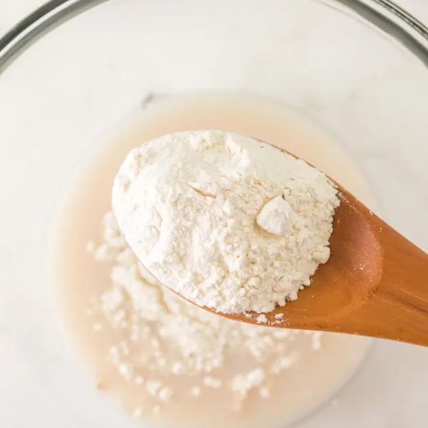 A wooden spoon adding flour to a wet yeast mixture in a bowl. Hostess At Heart