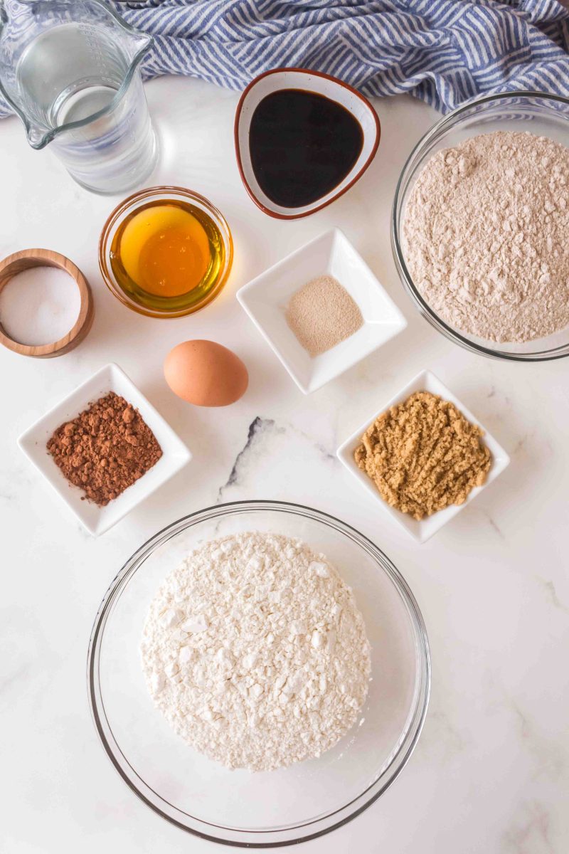 Ingredients needed to make molasses bread rolls including flour, honey, molasses, yeast, salt, brown sugar, water, cocoa, and an egg.