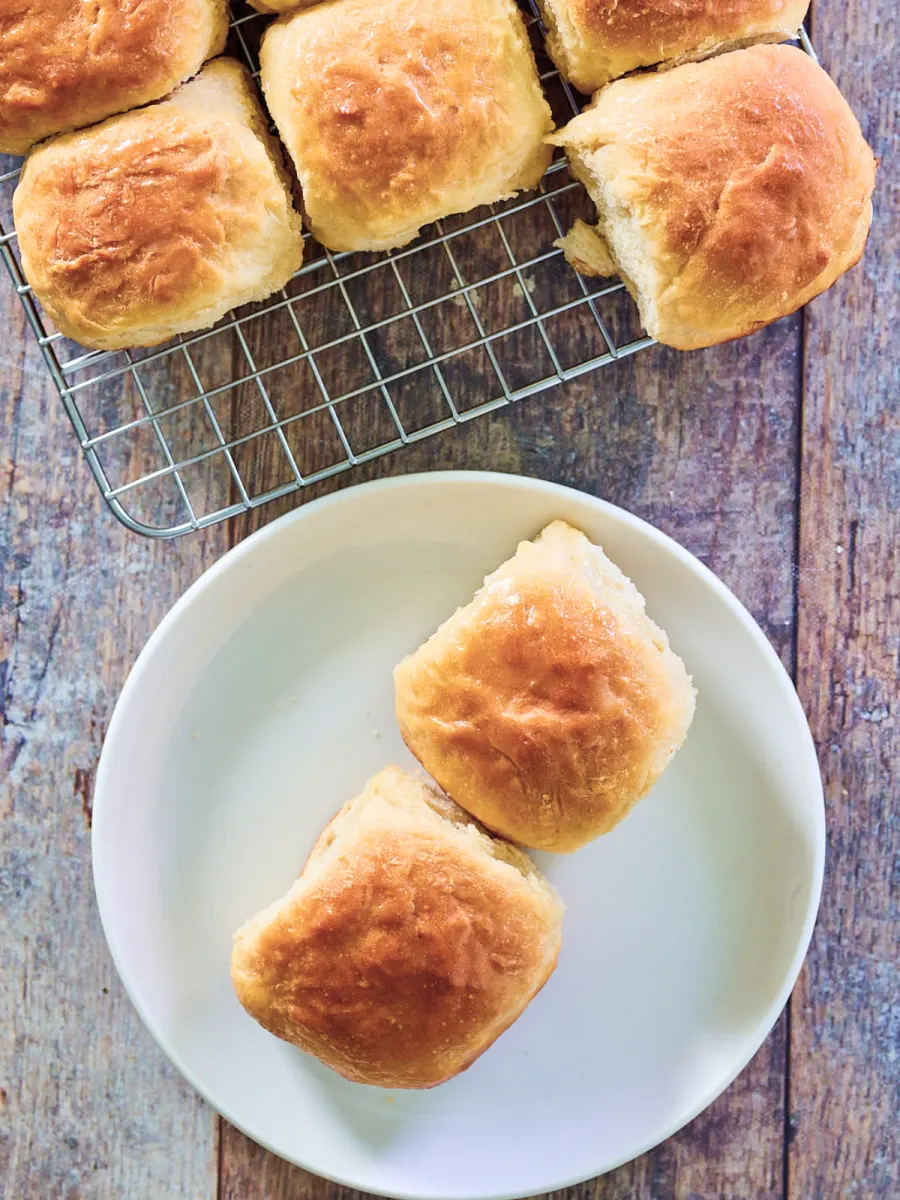 Top down view of two sweet hawian rolls on a plate with the rest of the rolls in the background on a cooling rack. Hostess At Heart