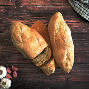 Two loaves of Garlic Batard Bread sitting on a wooden backdrop. One of the loaves is sliced. Hostess At Heart