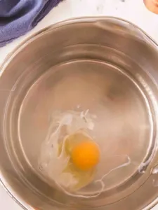 An egg just dropped into hot water for poaching. The egg white is just beginning to cook and the yolk remains bright yellow - Hostess At Heart