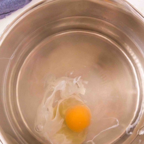 An egg just dropped into hot water for poaching. The egg white is just beginning to cook and the yolk remains bright yellow - Hostess At Heart
