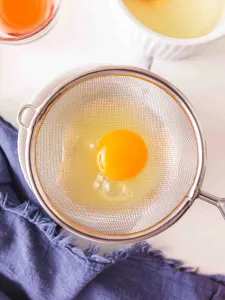 An egg sitting in a fine mesh strainer draining the watery whites from it before poaching - Hostess At Heart