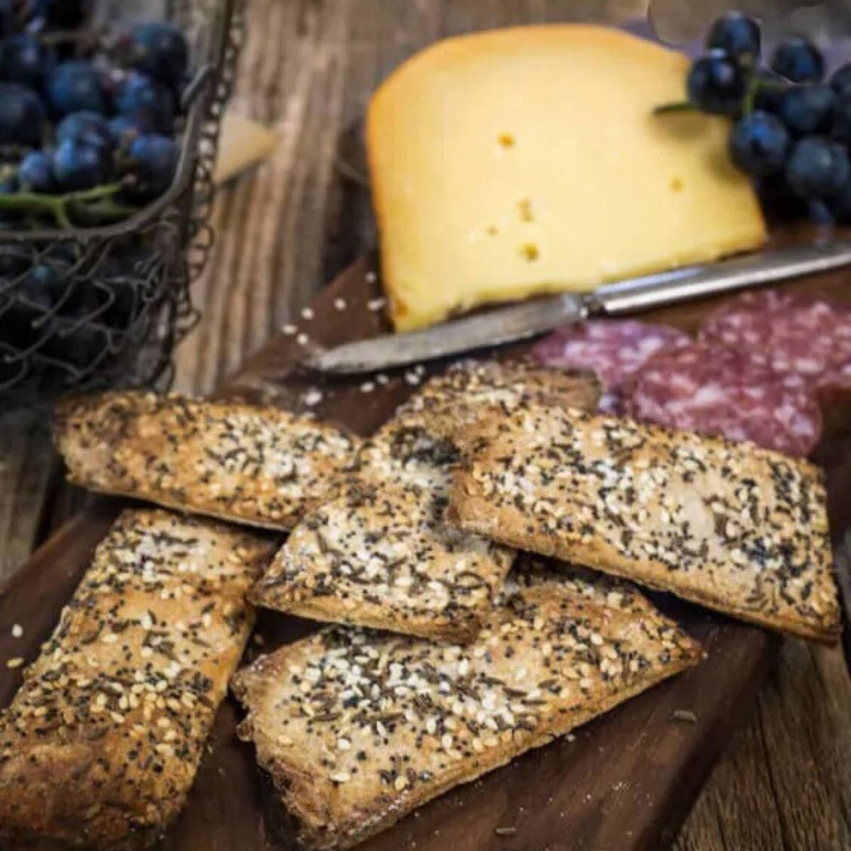 A pile of seeded crackers sitting on a board with meats and cheeses. Hostess At Heart