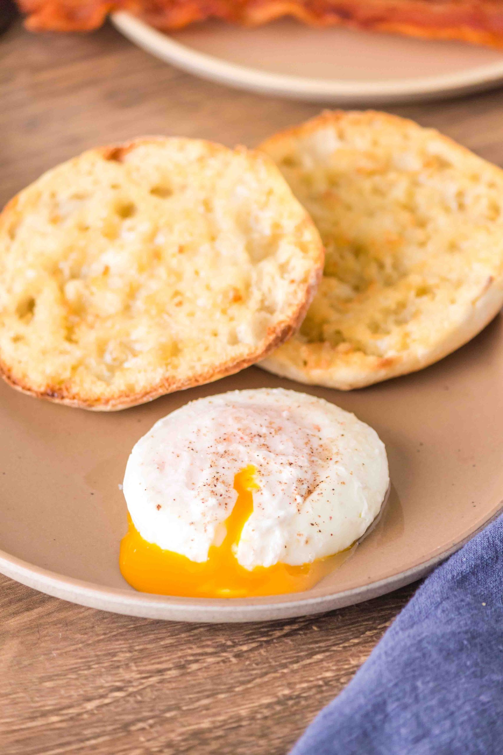 A poached egg on a plate that's been cut and has the yolk drizzling out in front of a split and toasted English Muffin