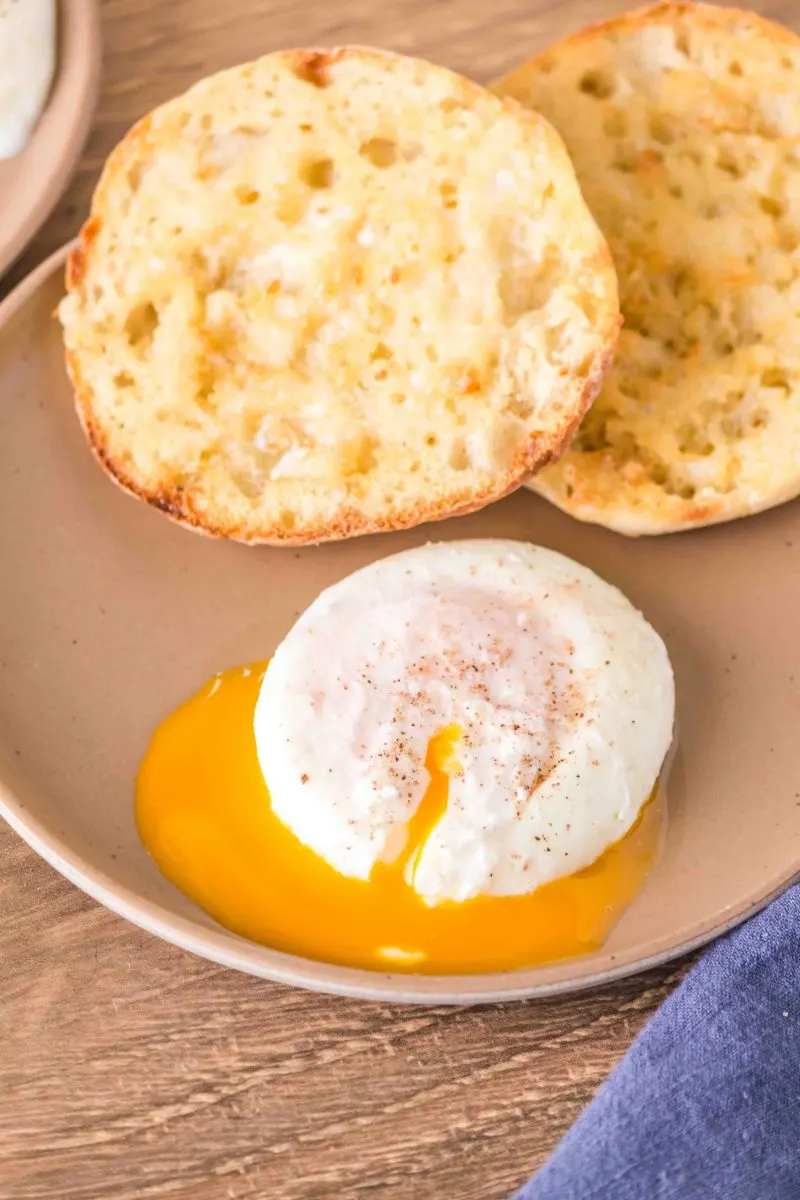 A plate holding a poached egg that has been cut and yolk is draining from the egg white next to a toasted English Muffin - Hostess At Heart
