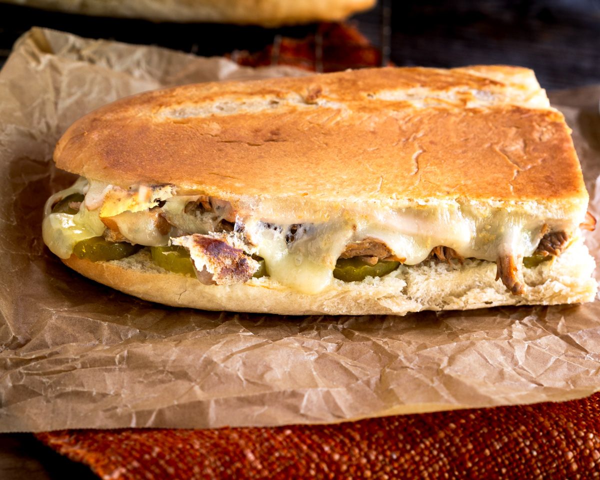 Sideview of a toasted Cubano sandwich showing melted cheese, shredded pork, ham and slices of pickles inside a toasted and pressed Cuban bread.
