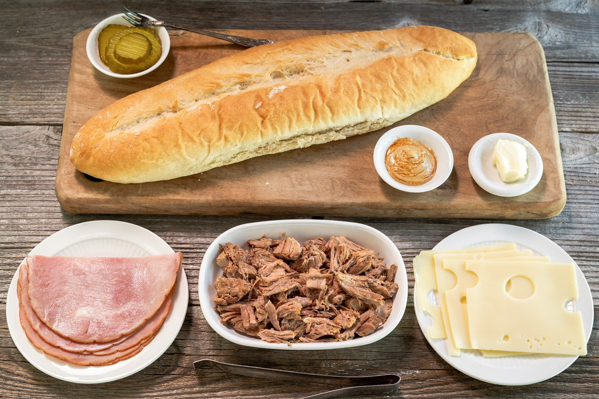 Ingredients used to make a Cuban Sandwich including Cuban Bread, Swiss Cheese, Cuban Mojo Pork, Sliced Ham, Pickles, Butter and mustard.