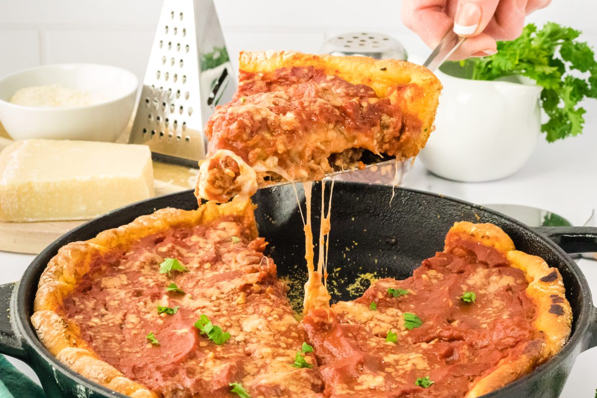 A slice of pizza with dripping melted cheese being lifted from a skillet holding the baked deep dish pizza - Hostess At Heart