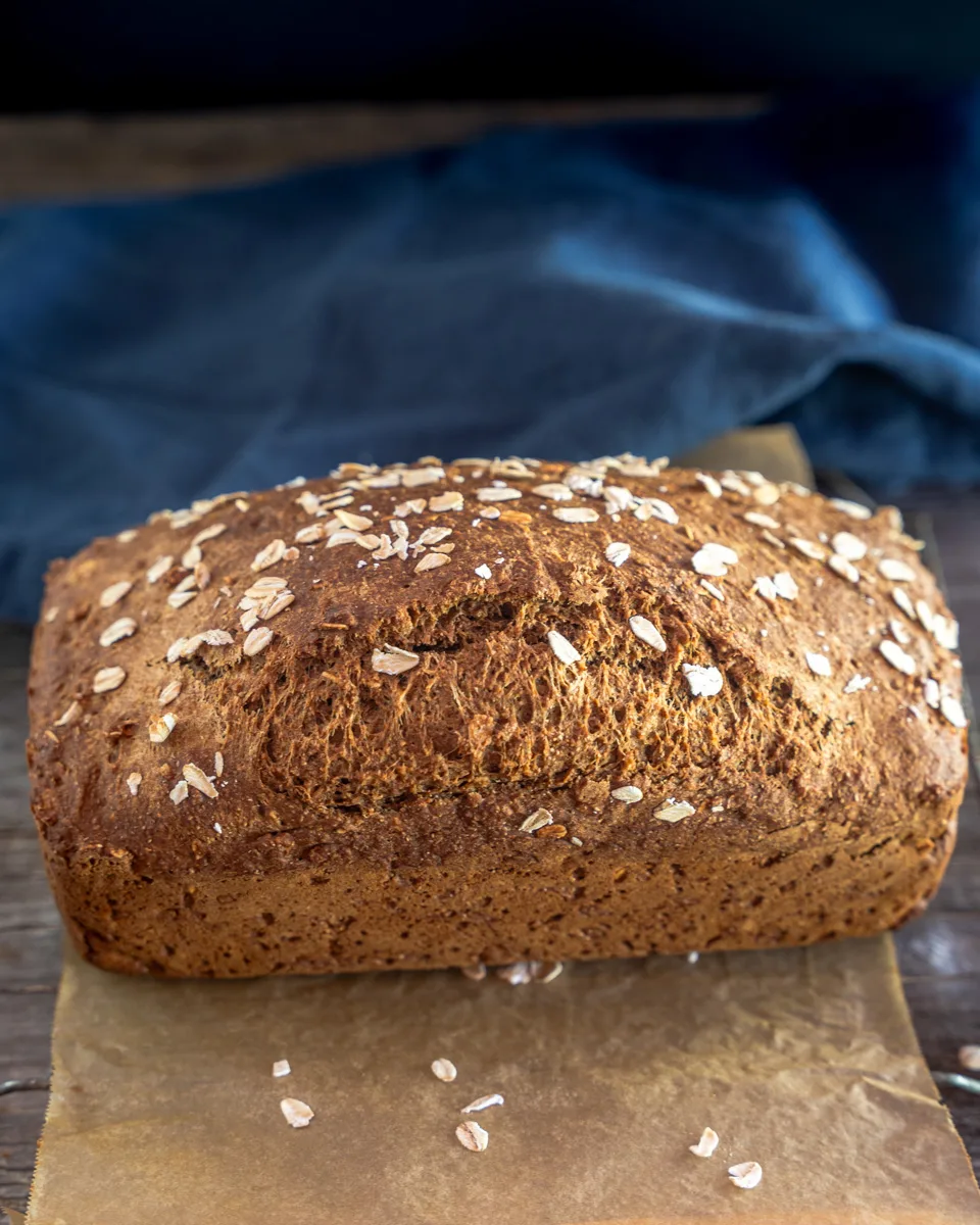 Sideview of a loaf of brown bread sprinkled with oats. The side edge is showing a slight blowout in the loaf.
