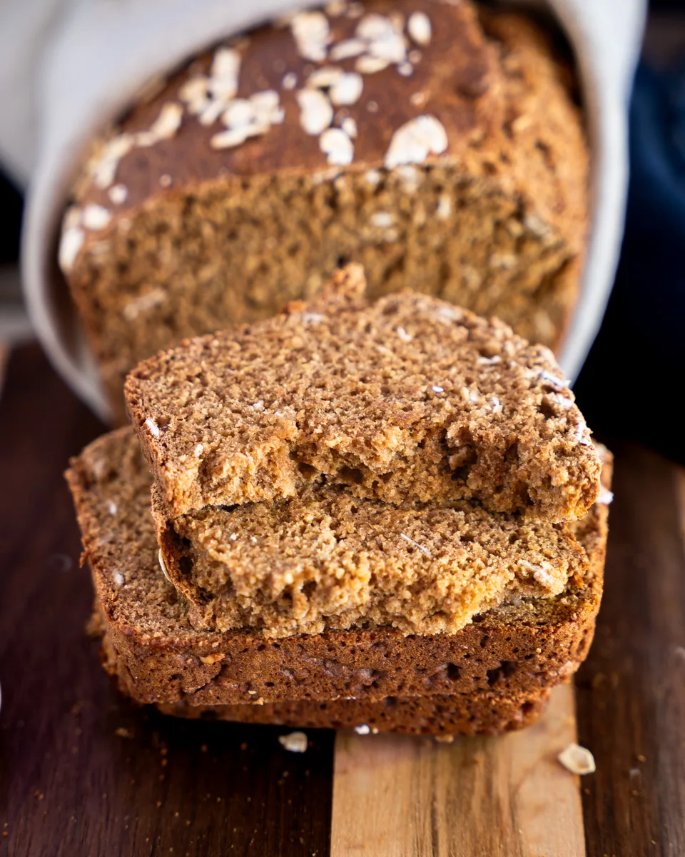 Front tableview of a loaf of brown bread with the first two slices laying in front of the loaf. The top slice is torn in half showing a soft crumb.