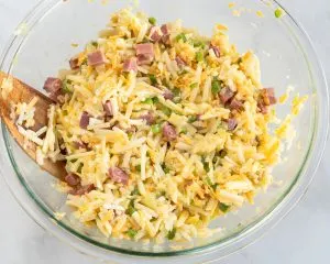 A bowl of combined corned beef breakfast casserole ingredients including shredded potatoes, cheese, egg green peppers, and onions. Hostess At Heart
