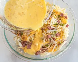 Egg mixture poured over shredded cheese, potatoes, diced corned beef, and vegetables in a mixing bowl. - Hostess At Heart