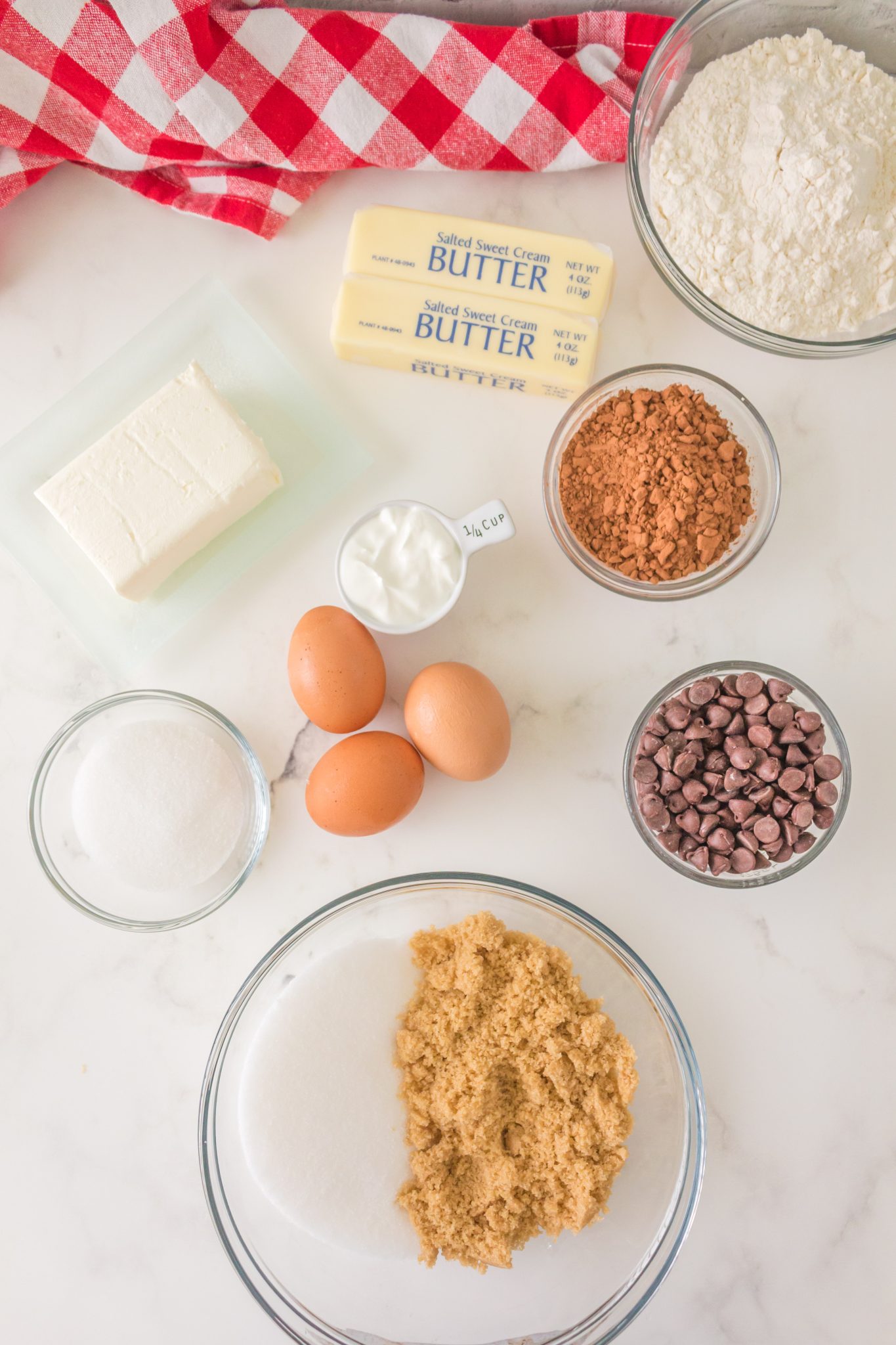 Top down view of ingredients: flour, sugar, butter, cream cheese, cocoa powder, eggs, chocolate chips, sugar, brown sugar, sour cream, vanilla extract. Hostess At Heart