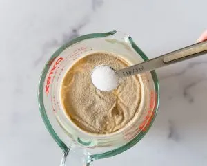 A measuring cup filled with yeast and water with a teaspoon of sugar being added in order to activate yeast. Hostess At Heart