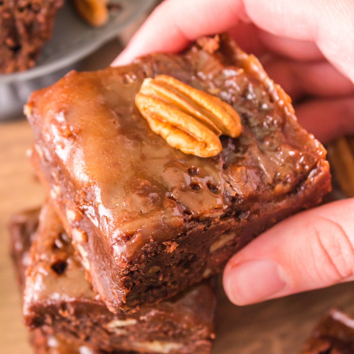 A hand holding a piece of Chocolate Turtle Brownie with a caramel topping and a pecan Hostess At Heart