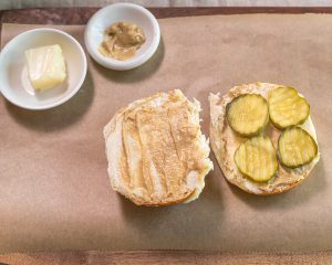 Top down view of Medianoche bread cut in half and buttered, slathered in mustard and topped with pickle slices - Hostess At Heart