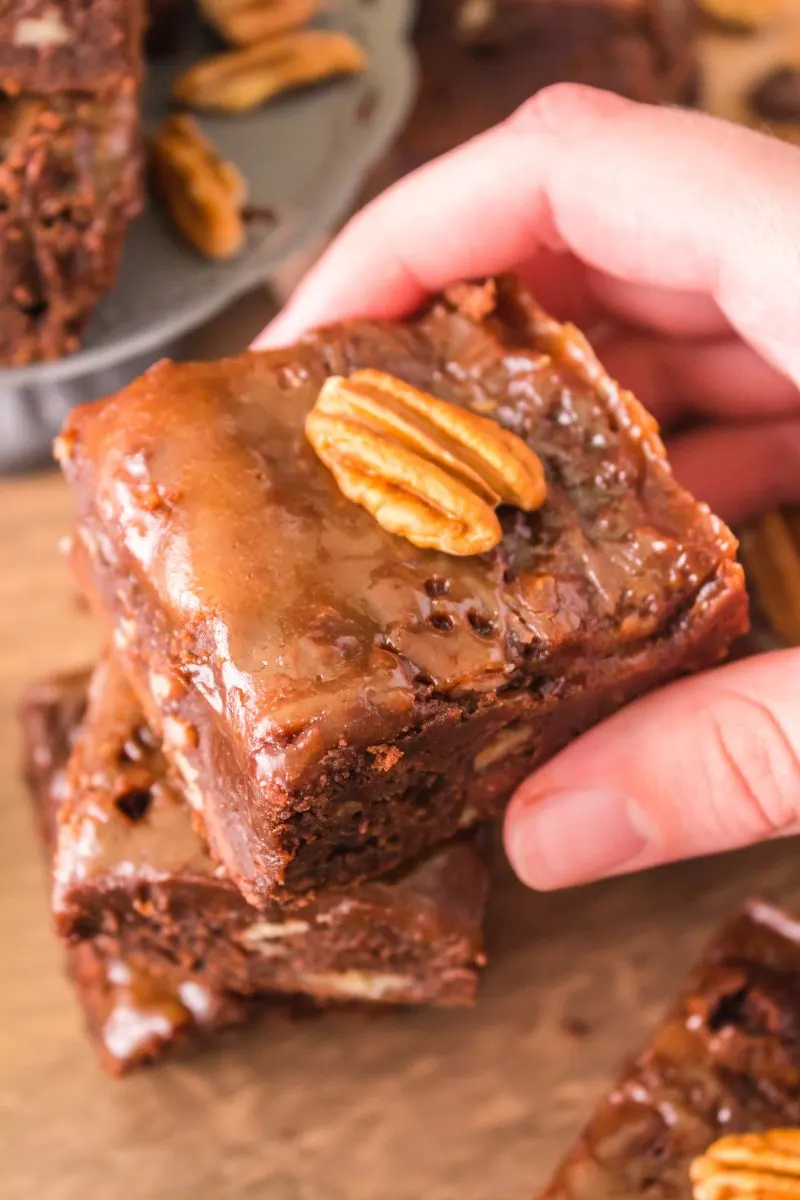 A hand holding a piece of Chocolate Turtle Brownie with a caramel topping and a pecan Hostess At Heart