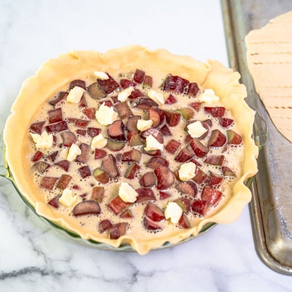Pieces of butter dotted over a rhubarb custard filling in a pie plate lined with pie crust - Hostess At Heart