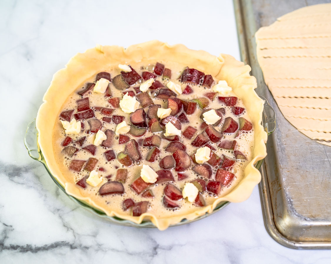 Pieces of butter dotted over a rhubarb custard filling in a pie plate lined with pie crust - Hostess At Heart