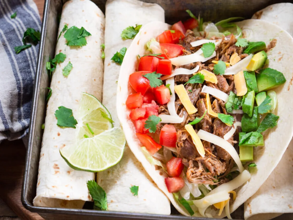 A pan filled with three rolled soft-shelled tacos with one open taco on top filled with insta pot shredded pork, diced tomatoes, avocado, and shredded cheese - Hostess At Heart