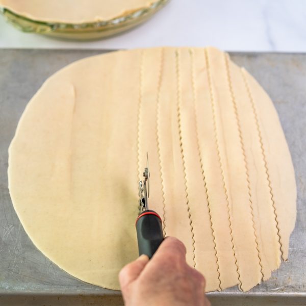 Rolled pie dough on a baking sheet being cut into strips with a ravioli cutter to make a lattice crust top - Hostess At Heart