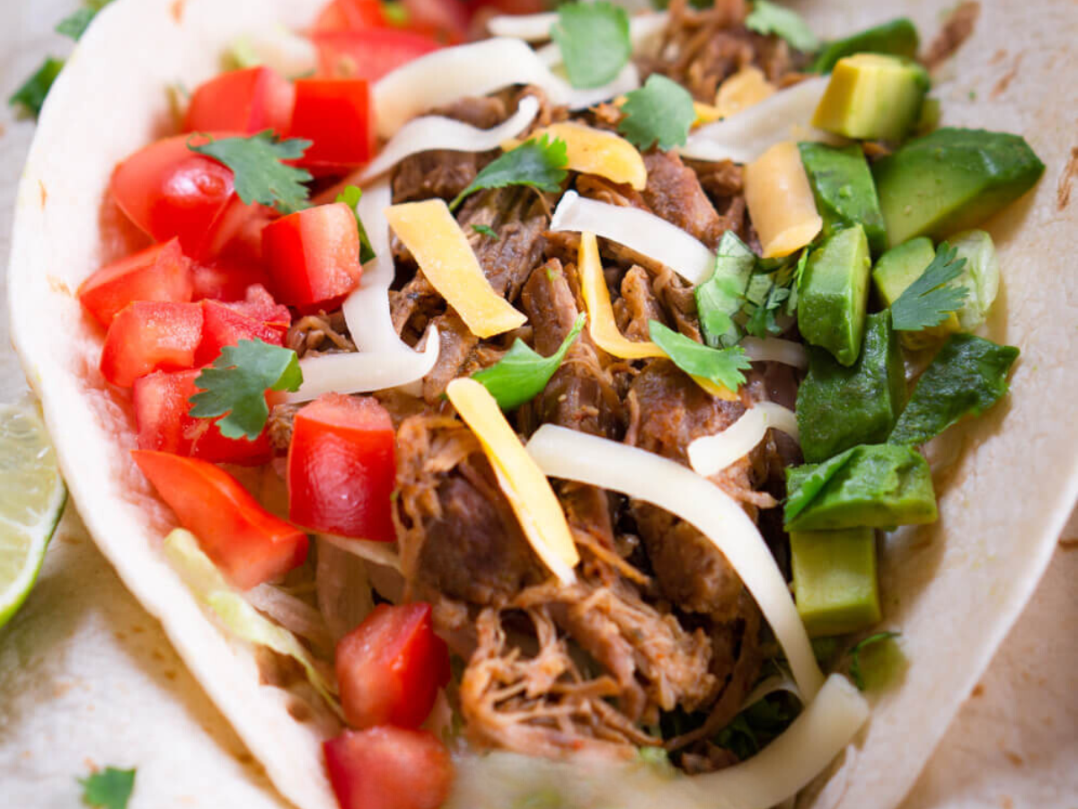 Closeup view of a taco shell filled with pulled pork taco meat, diced tomatoes, avocados, and shredded cheese.