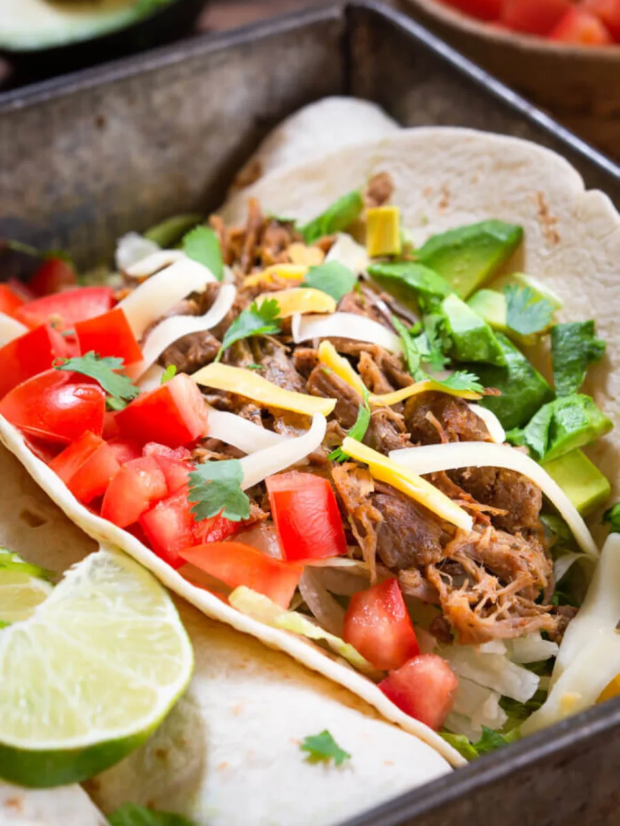 Side view of a soft shell taco tortilla filled with pulled pork and garnished with tomatoes, shredded cheese, and avocados. Hostess At Heart