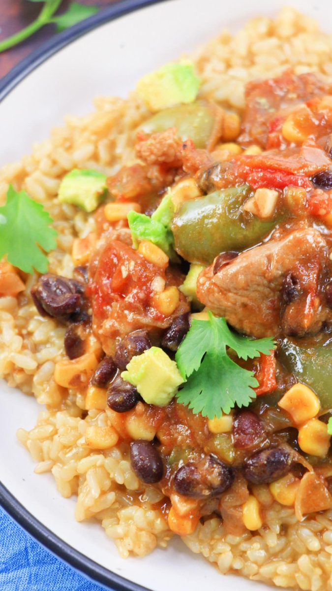 Close up view of a plate filled with soft cooked rice topped with a saucy stew filled with chunks of pork, black beans, onion, corn, tomatoes, and garnished with avocado and cilantro - Hostess At Heart