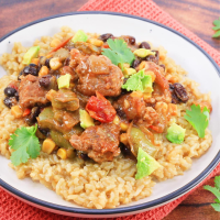 Angled view of a plate of tomatillo rice topped with chunks of pork stew meat, green chilies, black beans, corn, and tomatoes garnished with avocado and fresh cilantro - Hostess At Heart