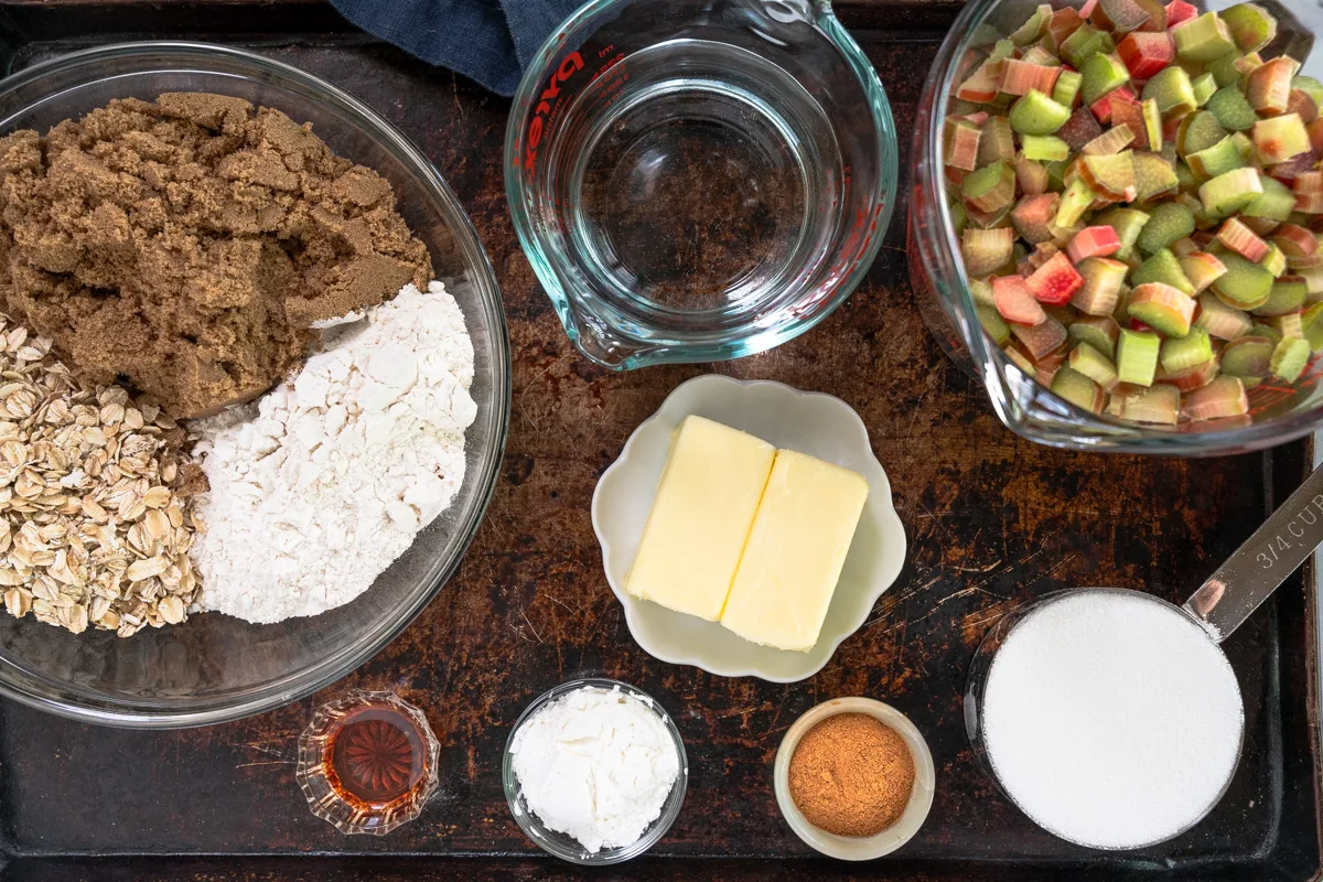 Top down view of ingredients used to make rhubarb crunch including brown and white sugar, flour, cinnamon, butter, vanilla, oats, rhubarb, cornstarch, and water.