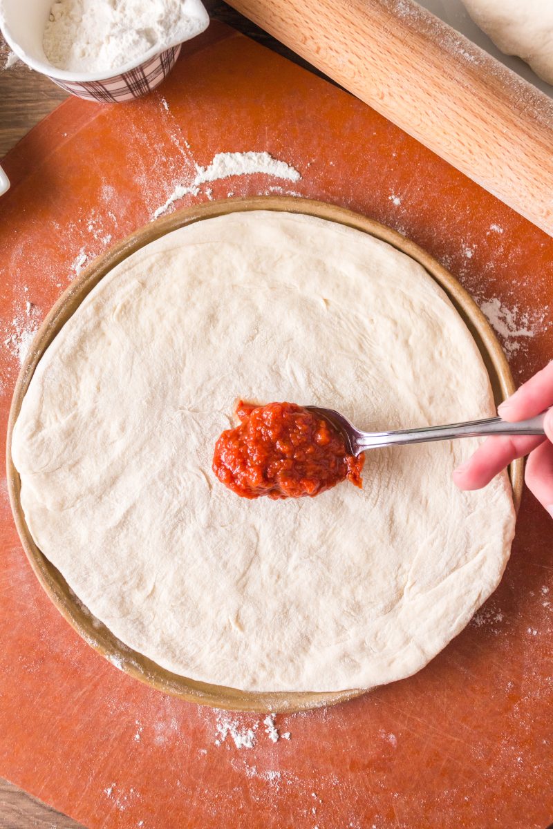 Top down view of a spoon of pizza sauce spread on a rolled out homemade sourdough pizza crust - Hostess At Heart