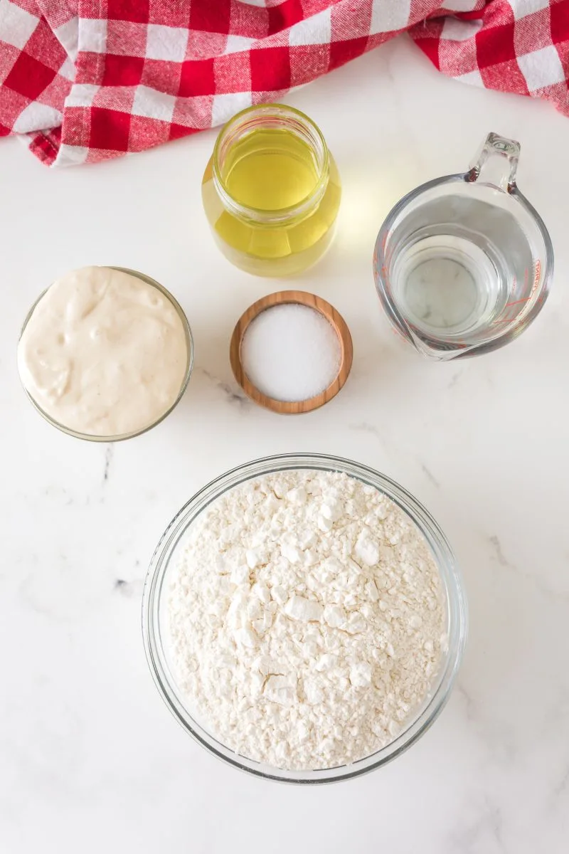 Top down view of ingredients used to make sourdough pizza crust including flour, olive oil, bubbly starter, salt and water
