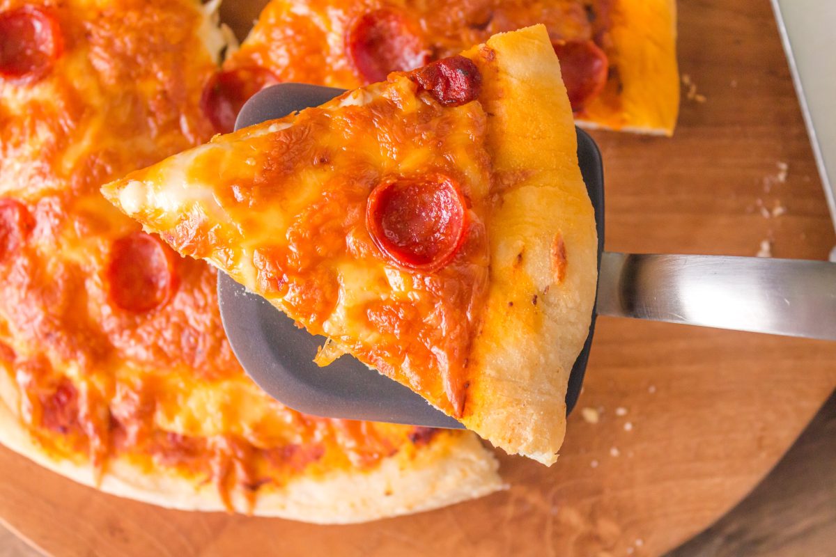 A slice of homemade pepperoni pizza made with sourdough pizza crust sitting on a spatula hovering over the pizza - Hostess At Heart