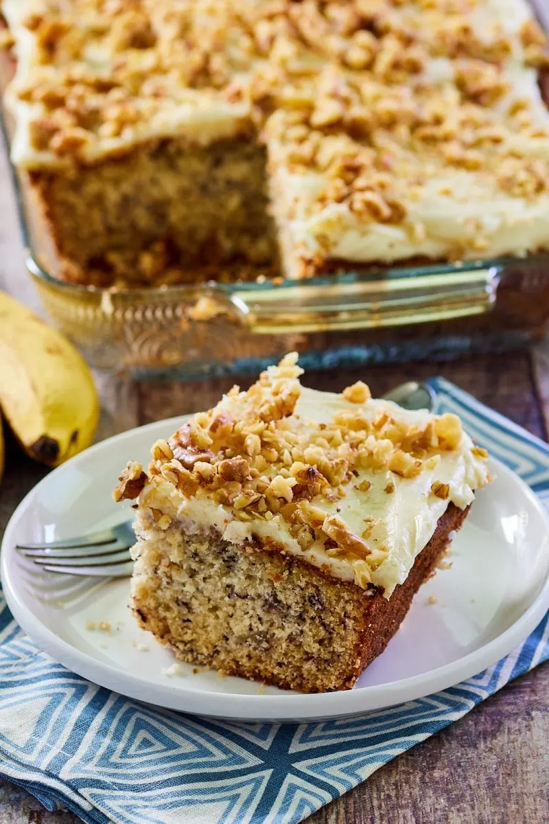 A sideview of a single piece of Banana Cake frosted and garnished with chopped walnuts - Hostess At Heart