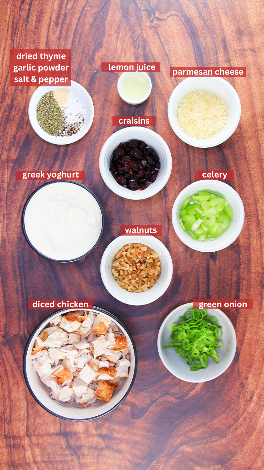 Top down view of bowls filled with the ingredients for chicken salad including chicken, celery, craisins, green onion, walnuts, greek yogurt, Parmesan cheese, lemon juice, thyme, garlic powder, salt, and pepper.