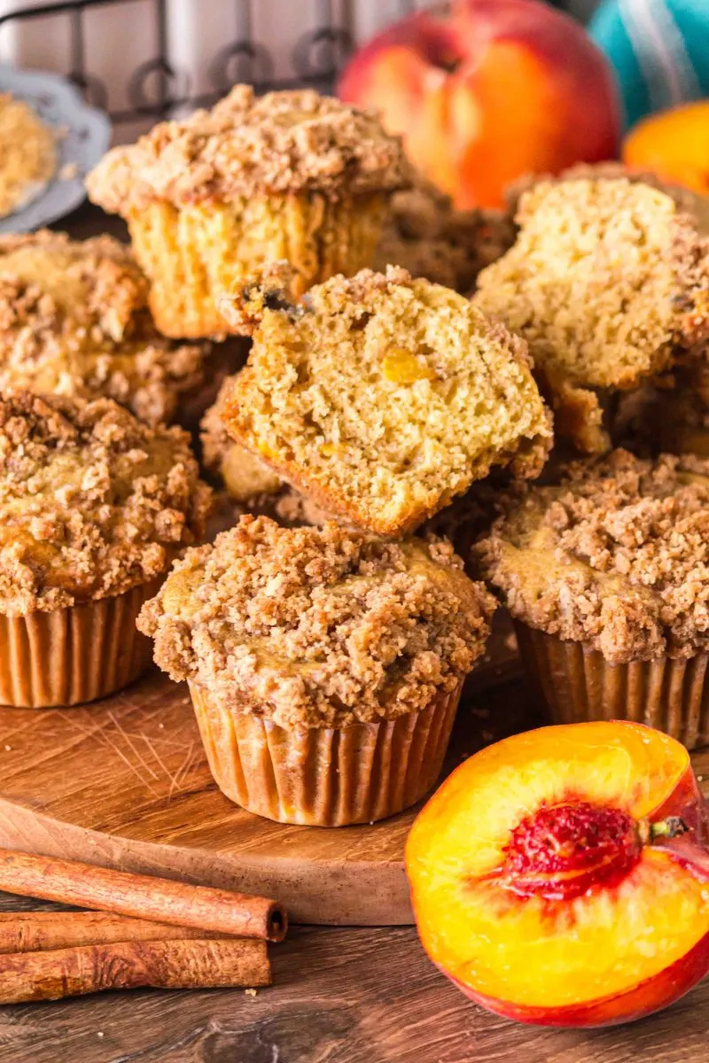 A pile of baked peach muffins topped with crumble with a half muffin on top showing a soft crumb - Hostess At Heart