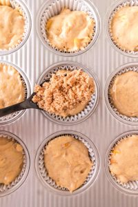 A spoon filled with streusel hovering over unbaked muffin batter in a cupcake pan. Hostess At Heart