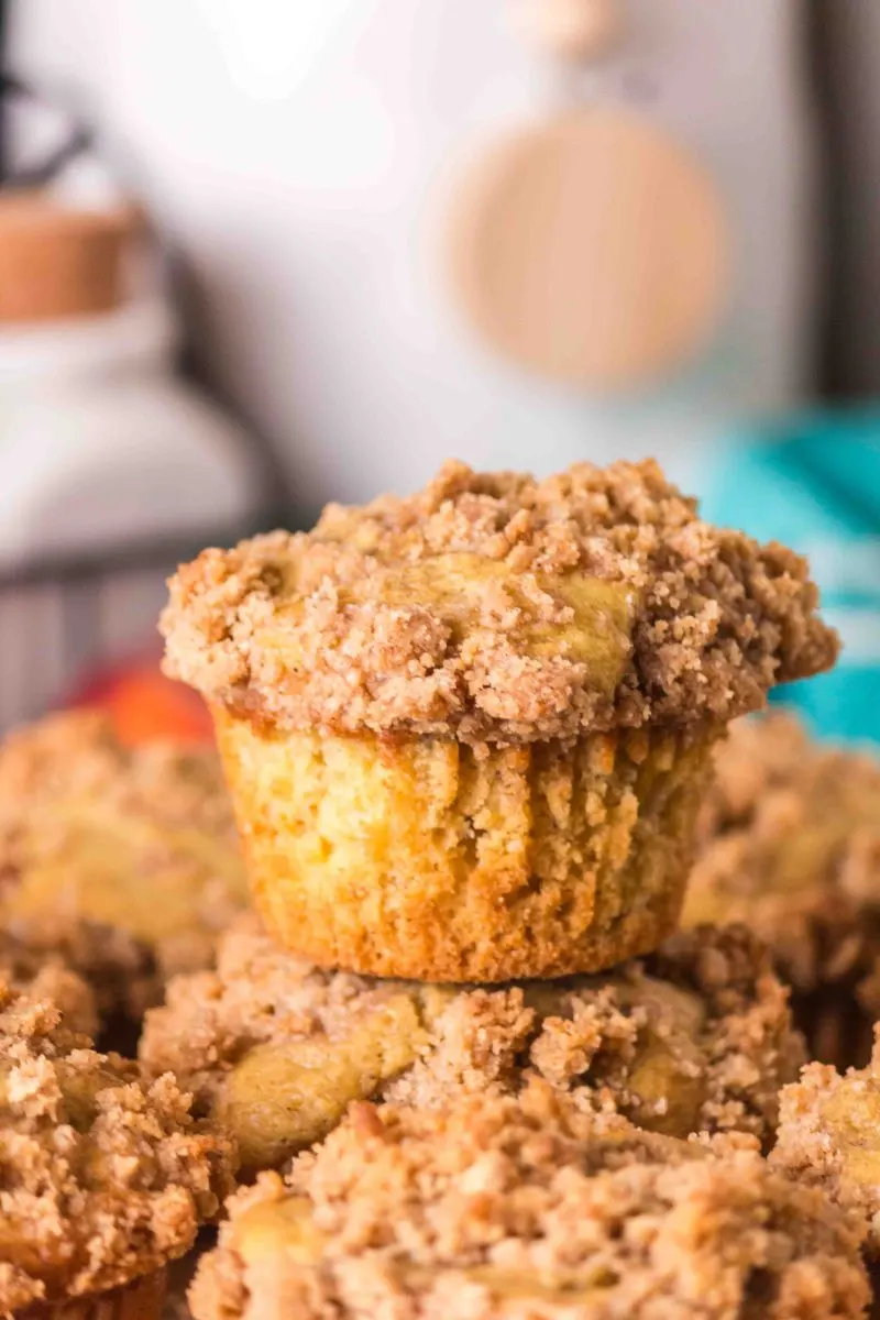 Sideview of a baked fresh peach muffin topped with crumble sitting on top of more peach muffins - Hostess At Heart