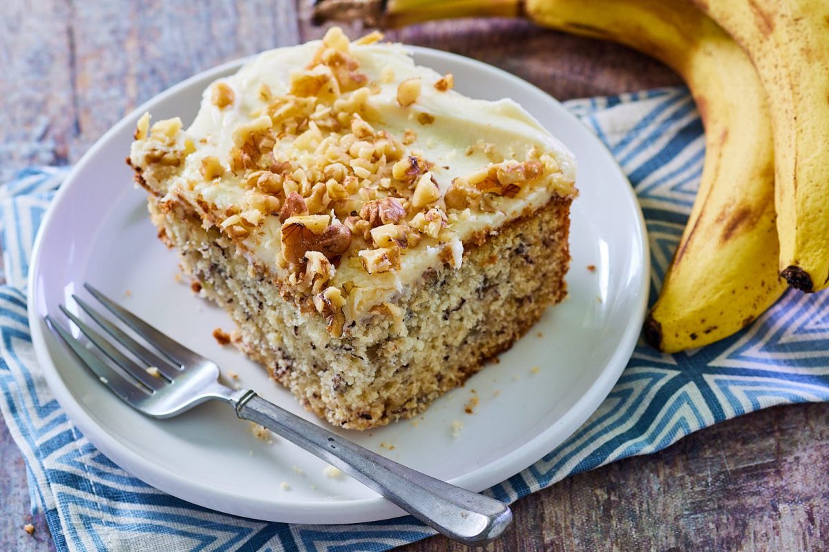 Close up view of a single slice of bannana cake topped with frosting and chopped nuts.