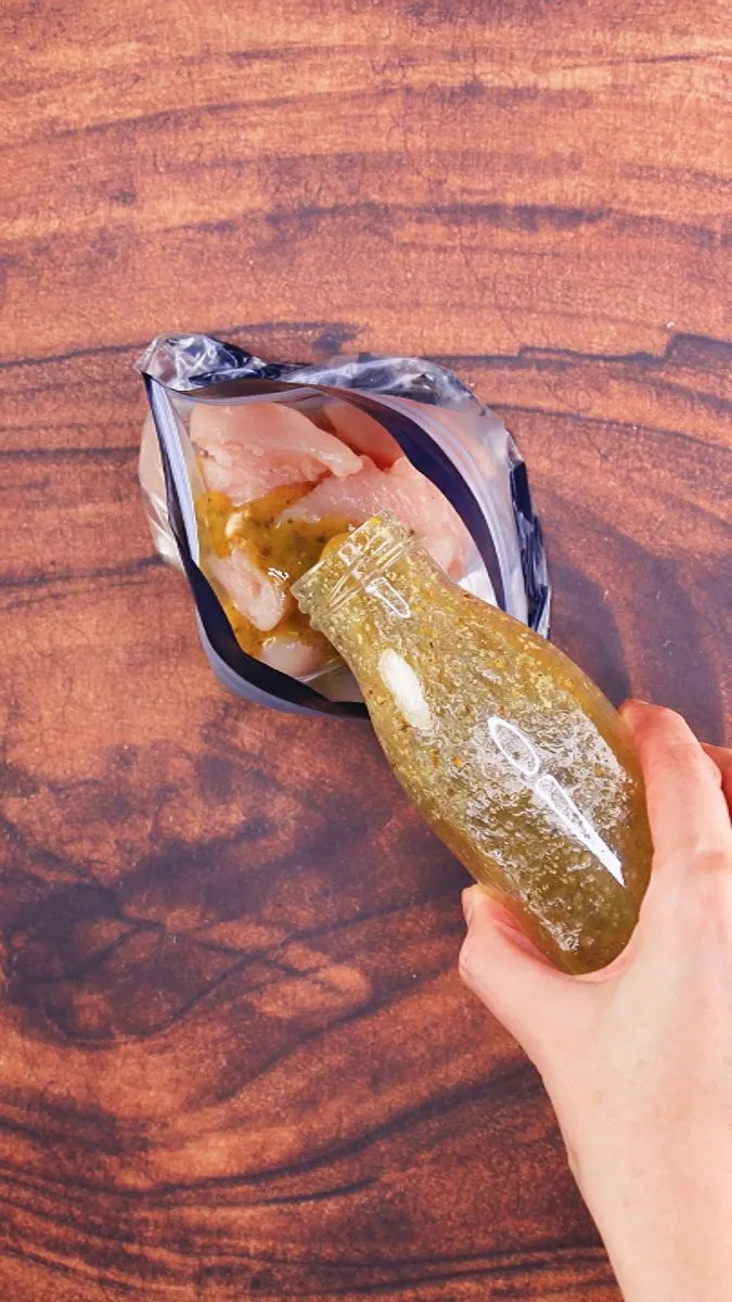 A glass bottle filled with a marinade being poured over chicken in a ziplock bag. Hostess At Heart