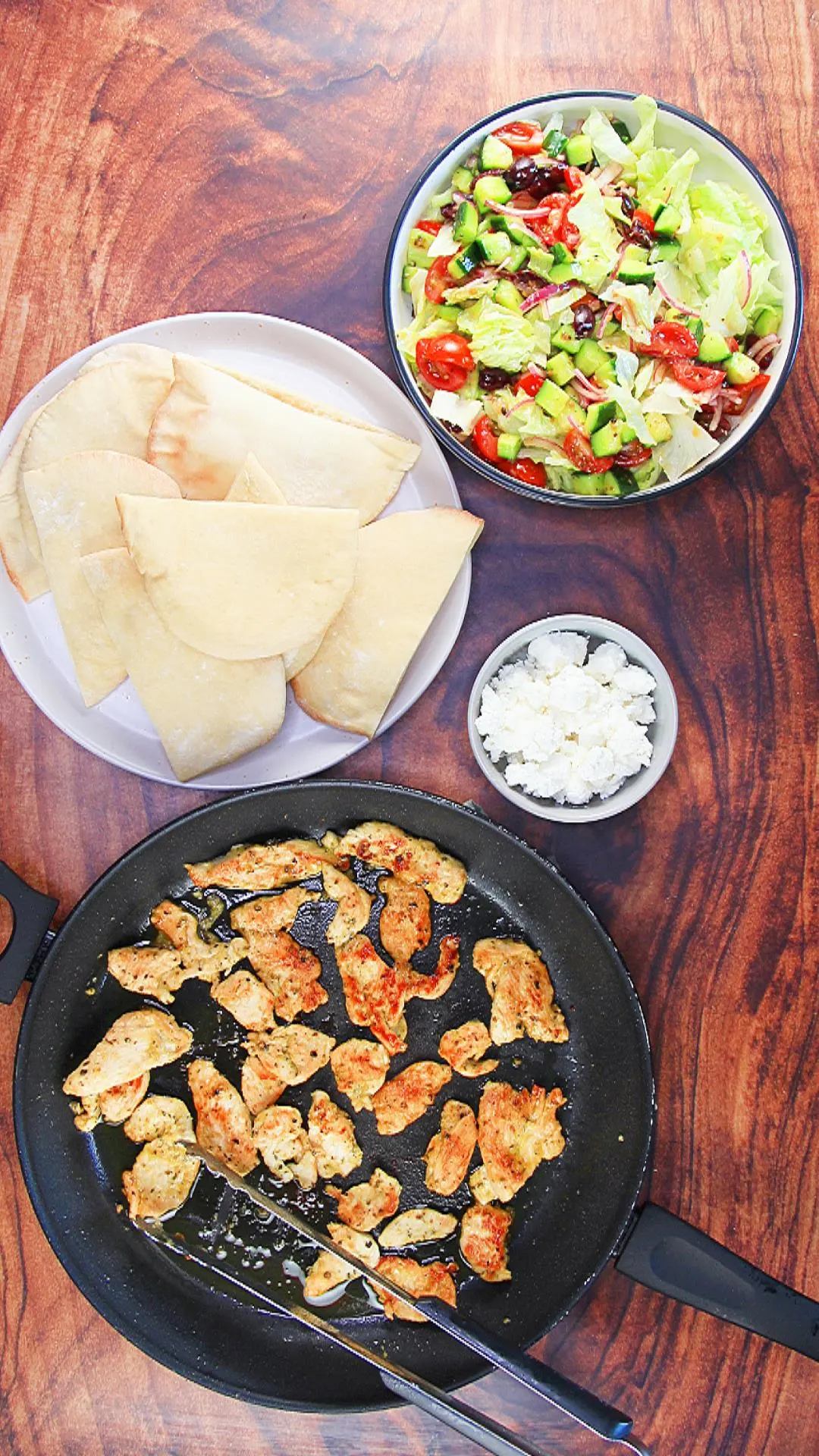 Top down view of ingredients used to make chicken gyros including sliced pita bread, browned chicken, lettuce, cucumbers, red onion, tomatoes and feta cheese.